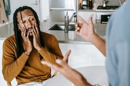 Man sitting at a table in a kitchen looks stressed with his face in his hands while another person gestures and speaks to him.