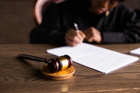 A judge’s gavel rests on a desk in the foreground while a person in the background writes on a document.