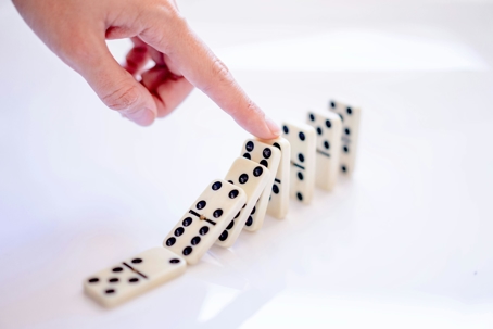 A hand touches a domino tile as a row of standing dominoes begins to fall in sequence on a white surface.