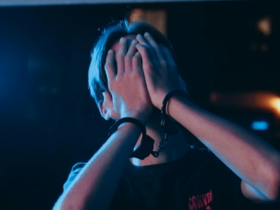 Person with hands covering their face while wearing handcuffs, lit with blue and red lighting in a dark setting.
