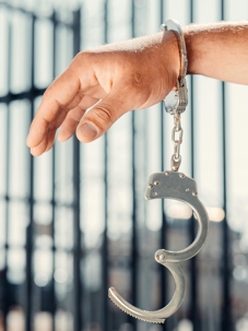 A close-up of a person’s wrist wearing an open metal handcuff, with vertical bars blurred in the background.