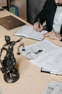 Person in a suit writing in a notebook at a desk with legal documents, a magnifying glass, a laptop, and a Lady Justice statue nearby.