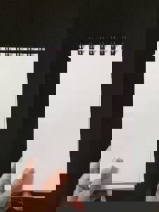 A hand holds a small blank spiral notebook with white pages against a dark background.