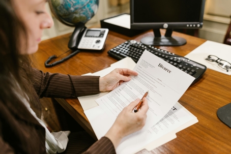 A person sits at a desk reviewing and marking a document titled “Divorce,” with a computer, phone, and office items visible in the background.