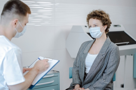 Masked doctor writing on a clipboard while speaking with a masked woman sitting in a medical examination room.