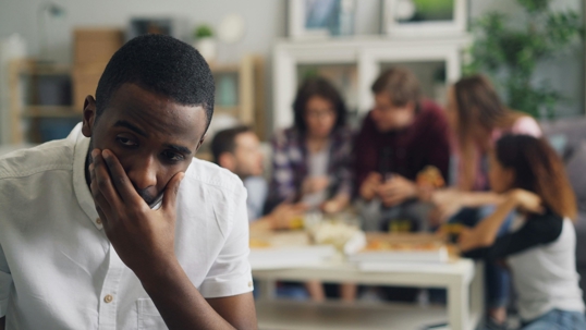 Man sitting in the foreground with his hand covering his mouth, looking worried, while a group of people socialize blurred in the background.