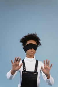 A person wearing a blindfold with hands outstretched, standing against a plain blue background.