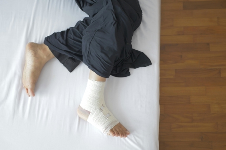 Person lying on a bed with one foot wrapped in a bandage, suggesting an injured ankle or foot.